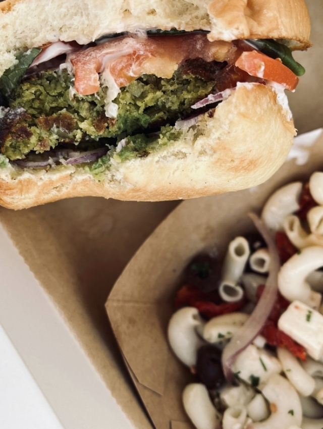 A close-up of a bitten falafel sandwich with tomato, greens, and sauce, next to a serving of macaroni salad with olives and red onions in a brown takeout container. Fearrington Village